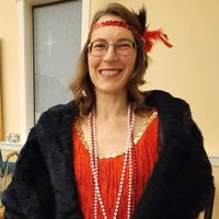 Allie, a smiling woman dressed in a 1920s flapper outfit with a red fringe dress, pearl necklaces, and a feathered headband stands indoors. She has glasses and drapes a black fur shawl over her shoulders.